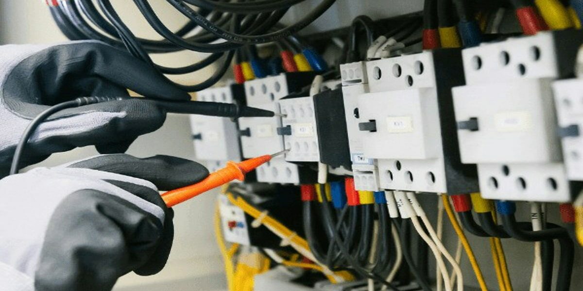 Électricien utilisant un multimètre pour contrôler un tableau électrique, avec câblage coloré et disjoncteurs visibles.